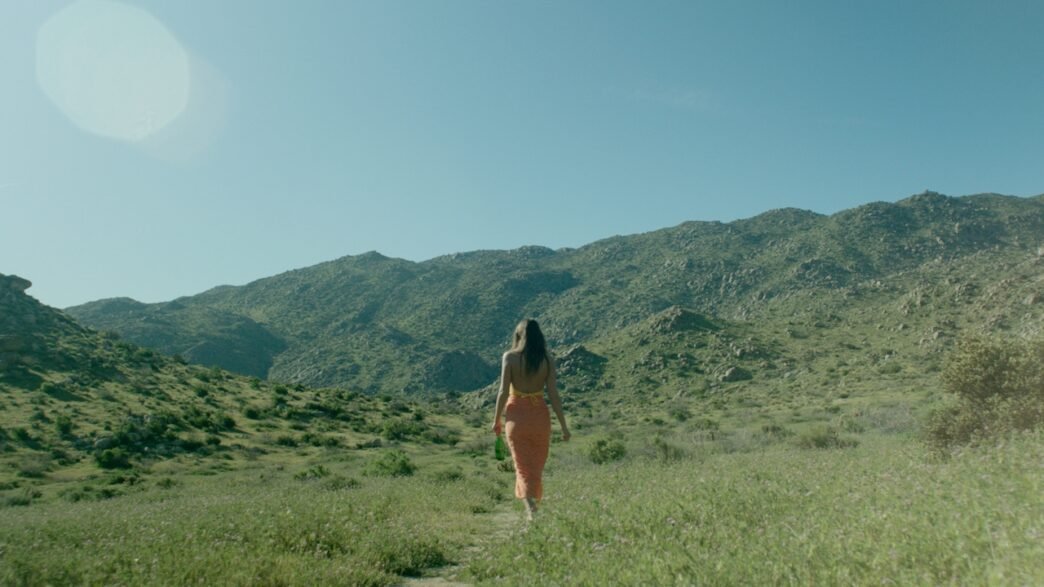 A women in a orange sundress and long brown hair walking in a grassy field with mountains in the background.