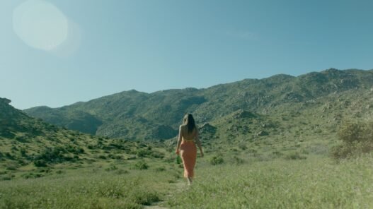 A women in a orange sundress and long brown hair walking in a grassy field with mountains in the background.