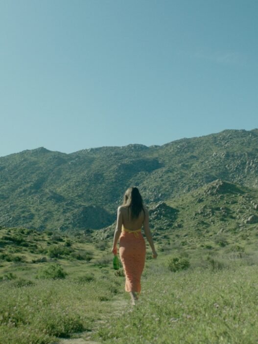 A women in a orange sundress and long brown hair walking in a grassy field with mountains in the background.