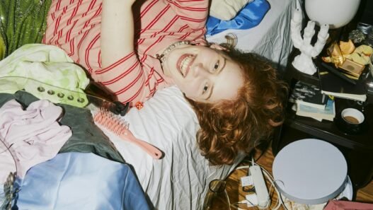 a young person laying on their bed looking playfully at their phone in a messy room