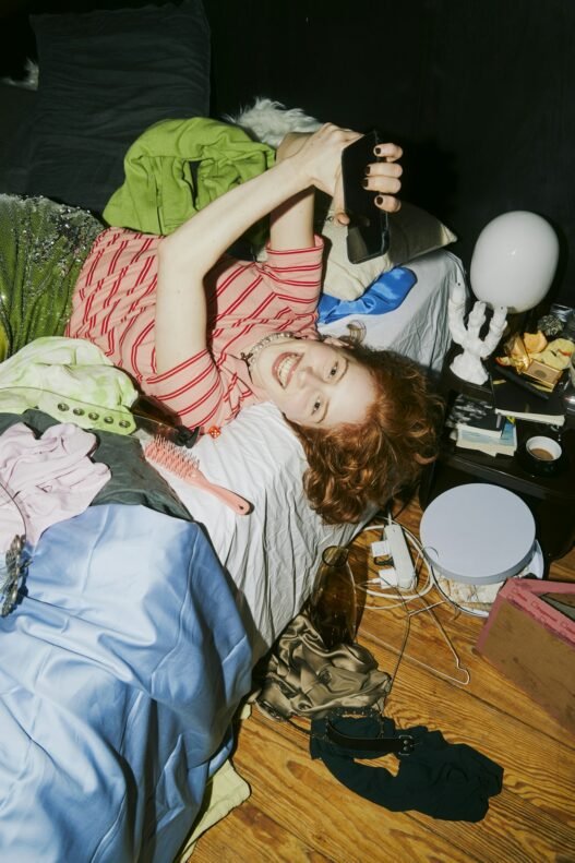 a young person laying on their bed looking playfully at their phone in a messy room