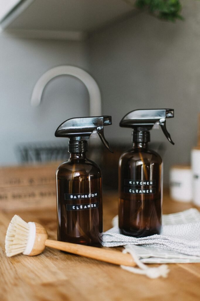 Two amber spray bottles with cleaning labels beside a wooden scrub brush.