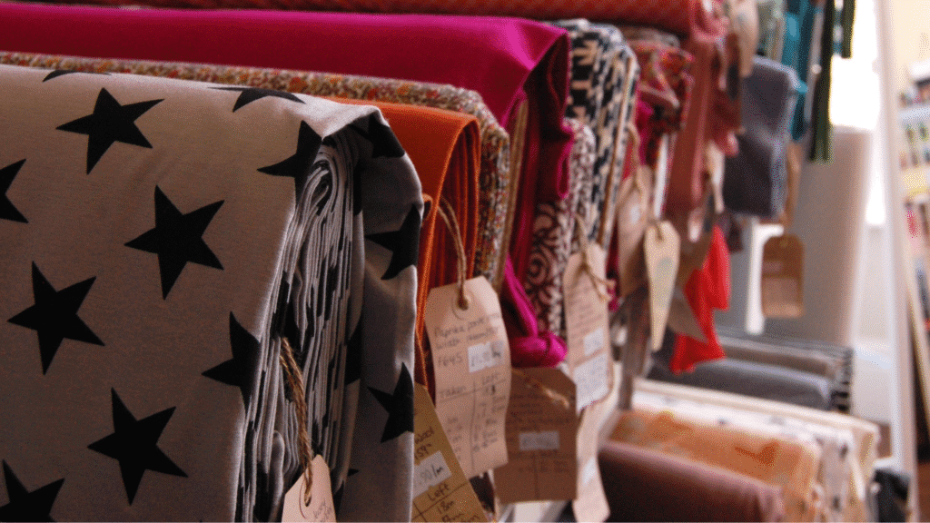 Bolts of fabric with fun patterns and bright colours sitting on a shelf in a dimply lit corner.