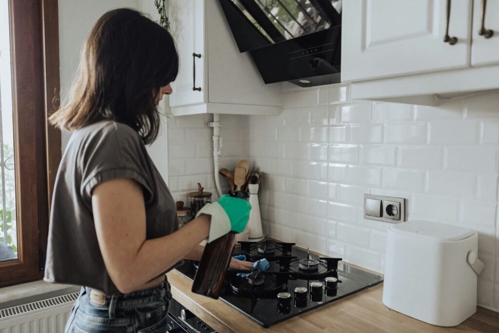 An image of a young person in a crop top with a spray bottle cleaning their stovetop.