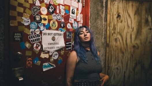 A young person with teal hair standing beside a double door at a punk rock bar covered in band stickers