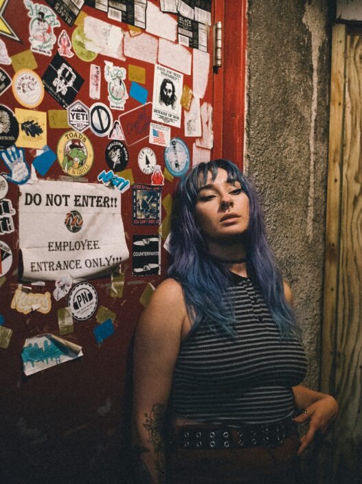 A young person with teal hair standing beside a double door at a punk rock bar covered in band stickers