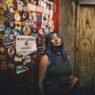 A young person with teal hair standing beside a double door at a punk rock bar covered in band stickers