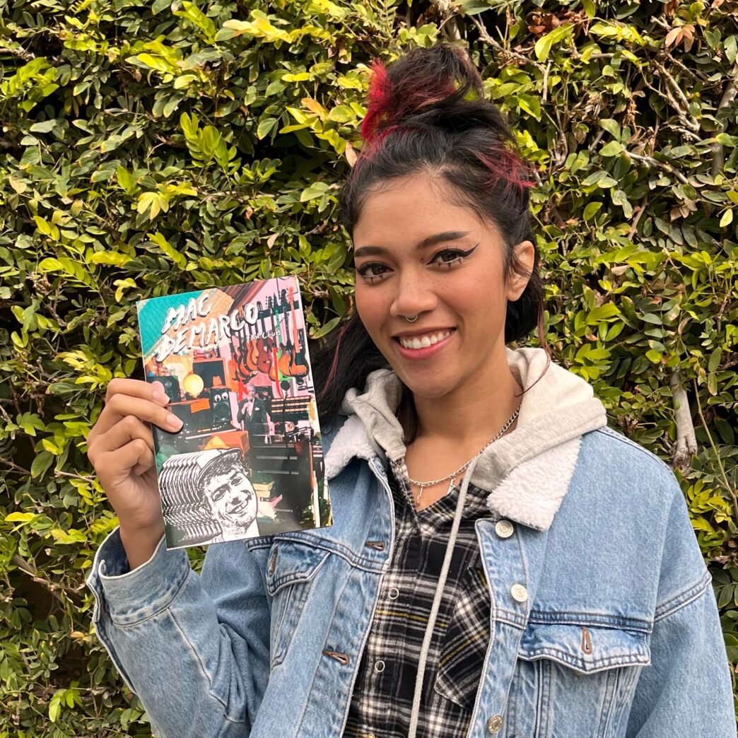 Bratty Bre wearing a demin jacket in front of a mossy green hedge holding a collection of zines.