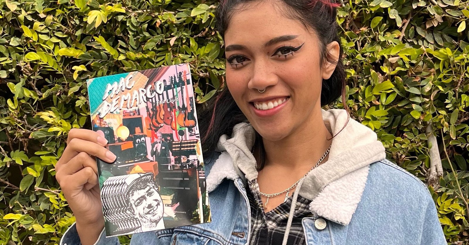 Bratty Bre wearing a demin jacket in front of a mossy green hedge holding a collection of zines.