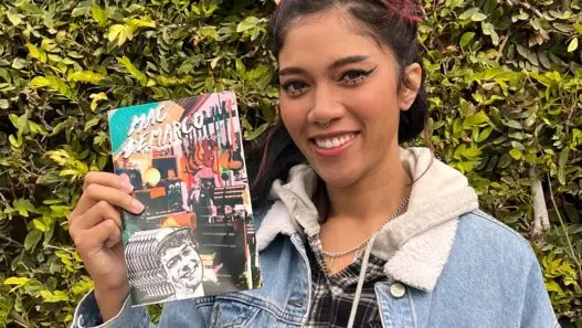 Bratty Bre wearing a demin jacket in front of a mossy green hedge holding a collection of zines.