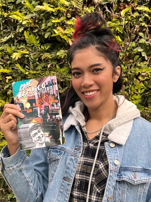 Bratty Bre wearing a demin jacket in front of a mossy green hedge holding a collection of zines.