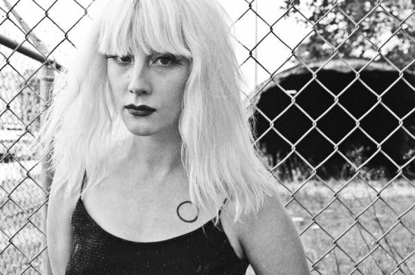 Mish Way in a black and white portrait standing in front of a chainlink fence.