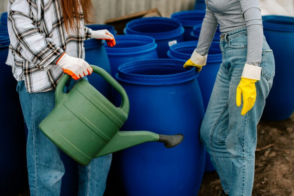 Two people standing in front of several blue water barrels, one carrying a large green plastic watering can.