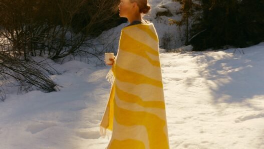 A woman walks through the snow with a white and yellow blanket draped over her shoulders and a hot mug in her hands.