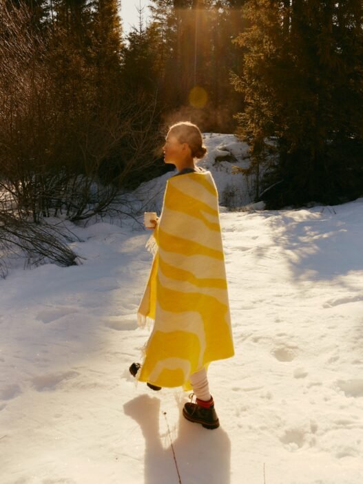 A woman walks through the snow with a white and yellow blanket draped over her shoulders and a hot mug in her hands.