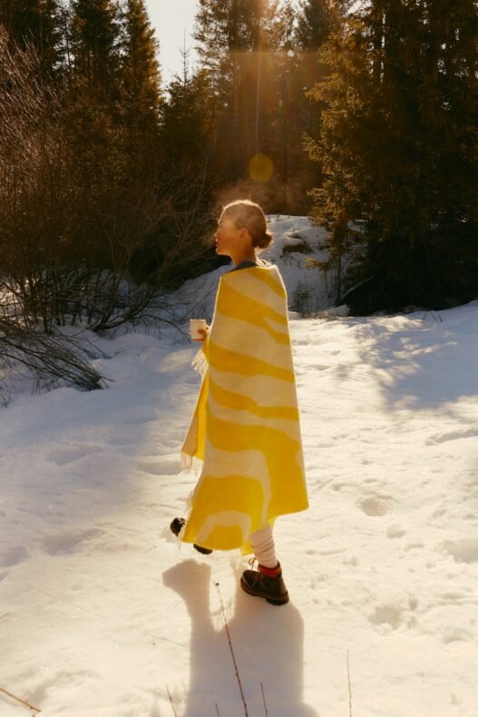 A woman walks through the snow with a white and yellow blanket draped over her shoulders and a hot mug in her hands.
