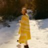 A woman walks through the snow with a white and yellow blanket draped over her shoulders and a hot mug in her hands.