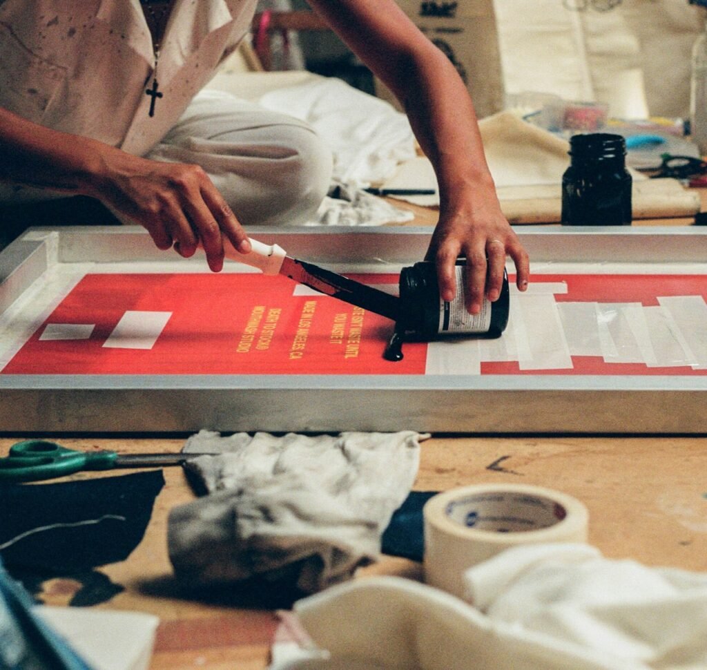 A person in a boiler suit sitting cross legged on the floor screen printing.
