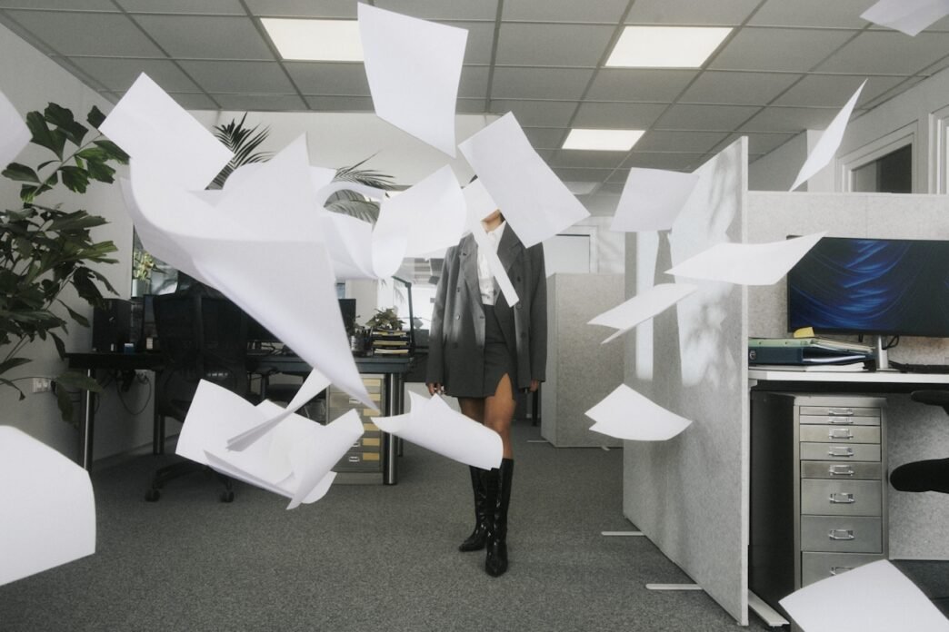 A woman standing in a blizzard of papers flying in the air in an office setting.