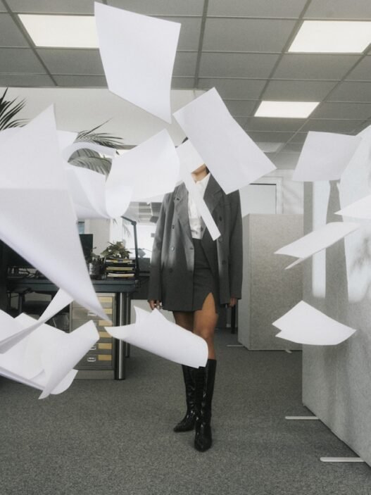 A woman standing in a blizzard of papers flying in the air in an office setting.