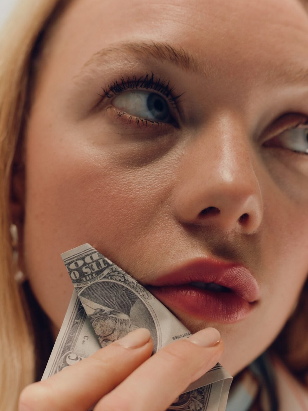 A blonde woman with a dollar bill held up against her cheek.