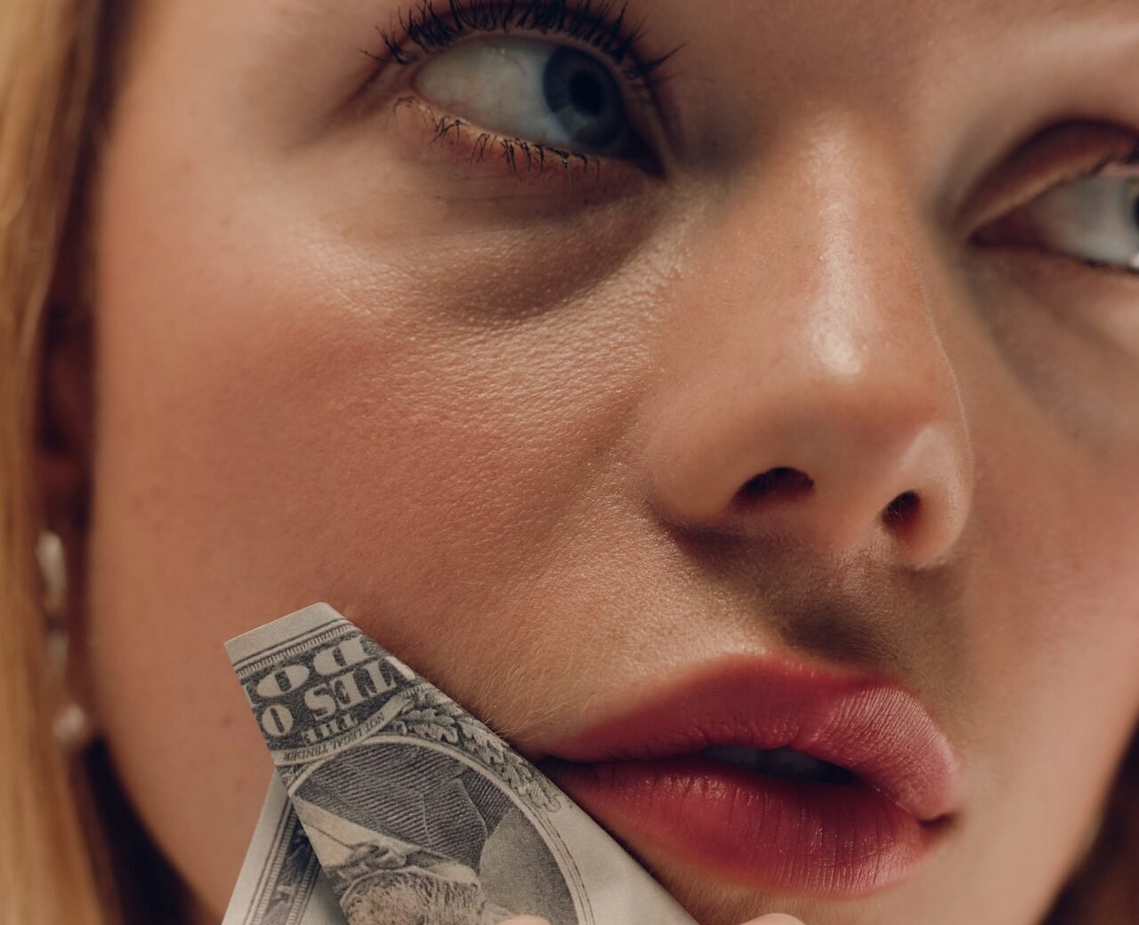 A blonde woman with a dollar bill held up against her cheek.