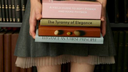 A women in a short skirt and knee high socks carrying a stack of books and standing in front of a bookcase.