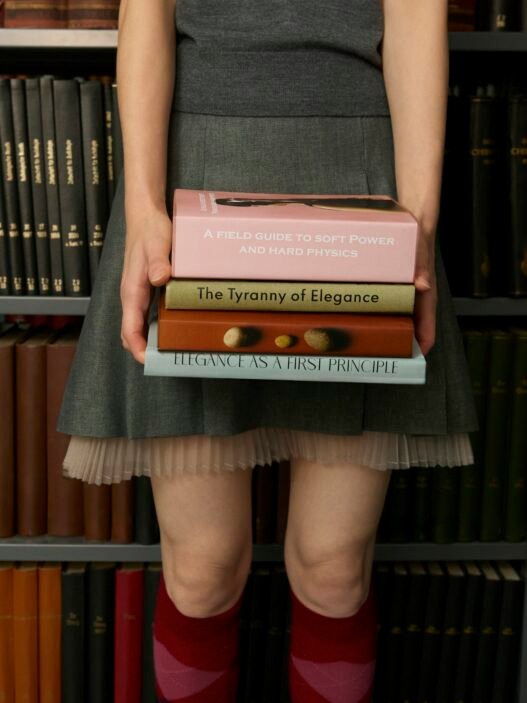 A women in a short skirt and knee high socks carrying a stack of books and standing in front of a bookcase.