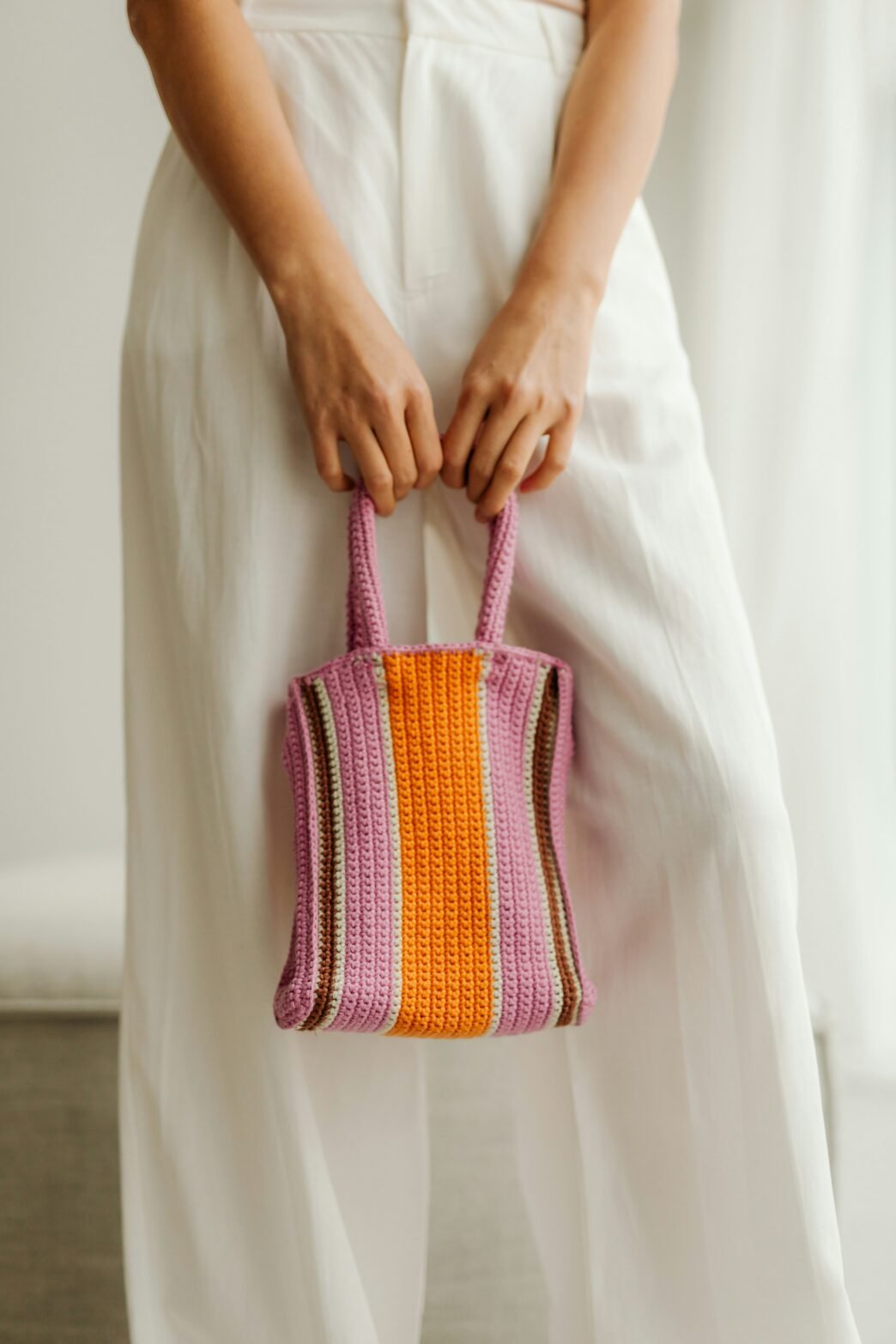 A woman in a long white dress holding an orange and purple striped crocheted purse.