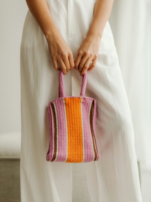 A woman in a long white dress holding an orange and purple striped crocheted purse.